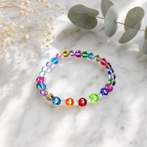 May include: A circular bracelet composed of translucent, multi-colored glass beads in shades of blue, purple, red, green, and yellow. Five of the beads are marked with the white numbers 1, 2, 3, 4, and 5 in a clear, sans-serif font.
