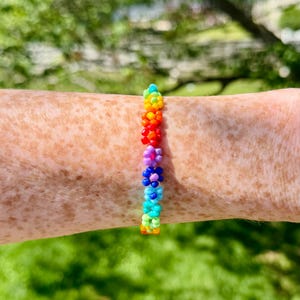 Pode incluir: Uma pulseira de missangas com uma série de pequenas flores multicoloridas dispostas em fila. Cada flor é construída a partir de minúsculas e vibrantes contas de vidro em tons de vermelho, laranja, amarelo, verde, azul e roxo.