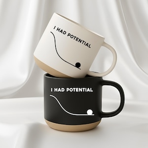 May include: Two ceramic mugs, one on top of the other. The top mug is white with a beige base, the bottom is black with a beige base. Both mugs feature the text "I HAD POTENTIAL" and a graphic of a ball rolling down a hill.