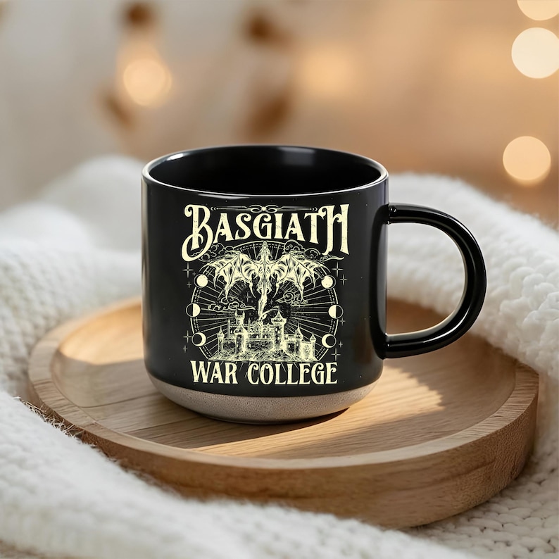 May include: Black ceramic mug with the text "BASGIATH WAR COLLEGE" in a gothic font. The mug features a detailed illustration of a winged creature, a castle, and celestial elements. The mug sits on a wooden tray.