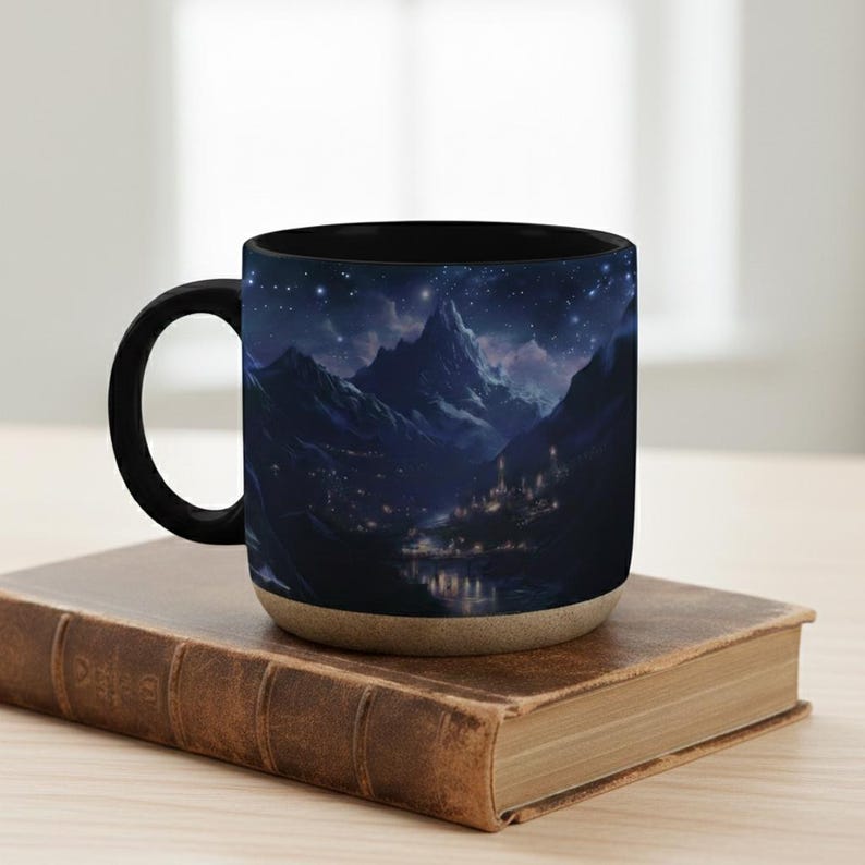 May include: A black coffee mug with a landscape design of mountains, a starry night sky, and a village. The mug has a black handle and rim, with a beige base. The mug is on a brown book.