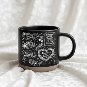 May include: Black ceramic mug with a tan base, featuring white illustrations and text. The design includes phrases like "Grumpy x Sunshine," "Just One More Chapter," and "Enemies to Lovers," along with book and floral motifs. The mug has a curved handle.