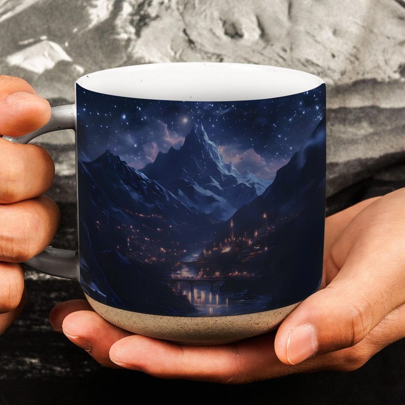 May include: A ceramic coffee mug with a mountain landscape design under a starry night sky. The image shows a mountain range, a village with lights, and a river. The mug has a white interior and a tan base.
