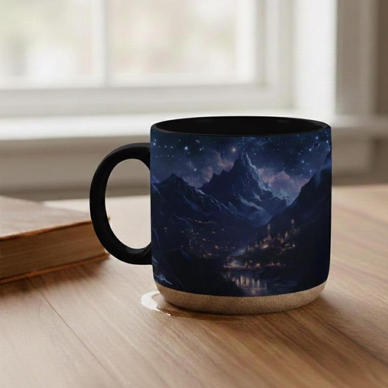 May include: A black coffee mug featuring a mountain landscape under a starry night sky. The mug has a black handle and rim, with a beige base. The design depicts a mountain range, a lake, and a village.