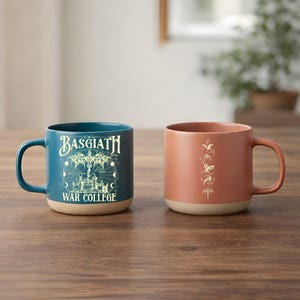 May include: Two ceramic mugs on a wooden table. One is teal with the text "Basgiath War College" and a graphic. The other is terracotta with a vertical design. Both mugs have a beige base and a handle.