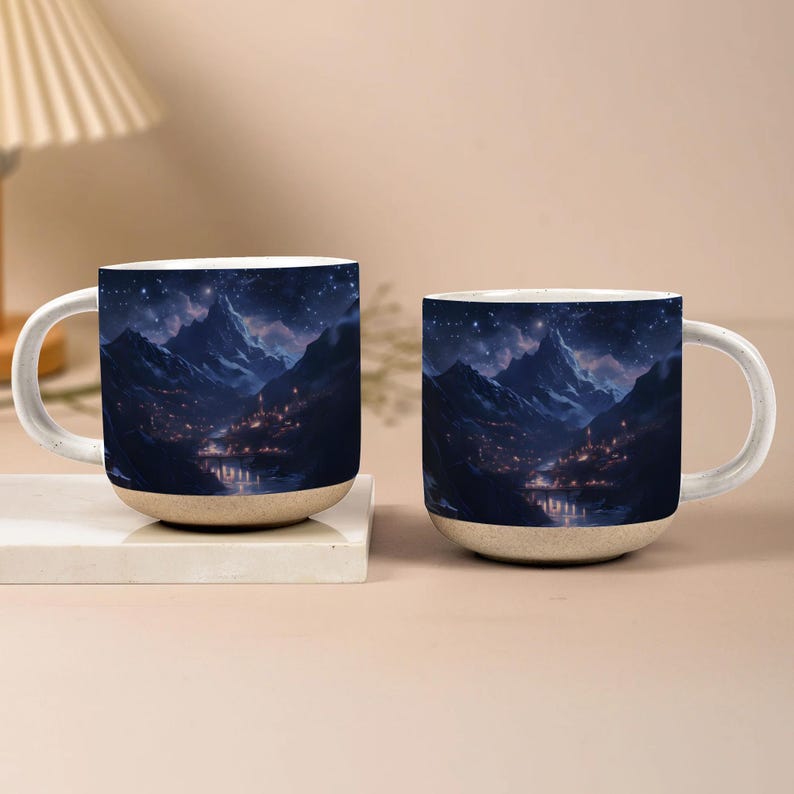 May include: Two ceramic mugs with a mountain landscape design. The mugs feature a night sky with stars, snow-capped mountains, and a town at the base of the mountains. The mugs have a white handle and a beige base.