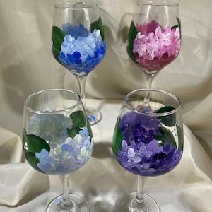 May include: Four wine glasses with hand-painted hydrangea designs. The glasses feature blue, purple, light blue, and pink hydrangeas.