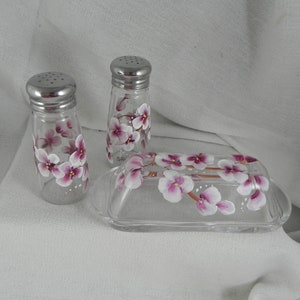 May include: A set of hand-painted glass salt and pepper shakers and a butter dish with a floral design. The shakers and dish are decorated with pink and white flowers.