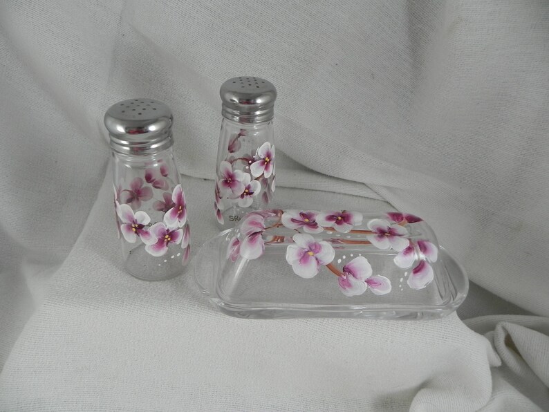May include: A set of hand-painted glass salt and pepper shakers and a butter dish with a floral design. The shakers and dish are decorated with pink and white flowers.