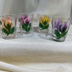 May include: Four clear glass shot glasses with hand-painted floral designs. Each glass features a different colored flower: peach, pink, yellow, and purple.