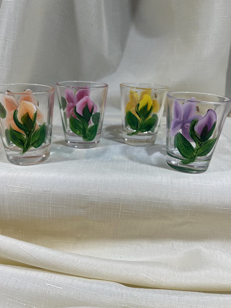 May include: Four clear glass shot glasses with hand-painted floral designs. Each glass features a different colored flower: peach, pink, yellow, and purple.