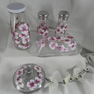 May include: Hand-painted glass kitchenware set featuring a floral design. The set includes a butter dish, salt and pepper shakers, and a jar with a lid. The flowers are pink and white.