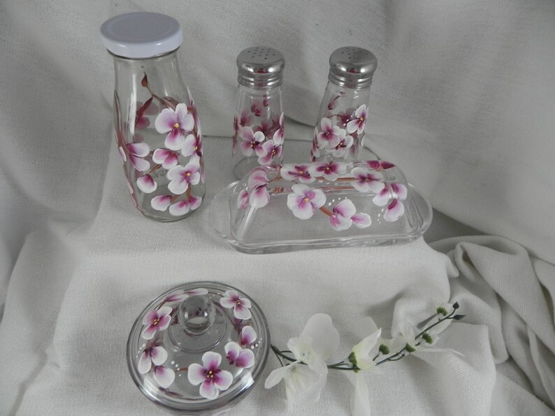 May include: Hand-painted glass kitchenware set featuring a floral design. The set includes a butter dish, salt and pepper shakers, and a jar with a lid. The flowers are pink and white.