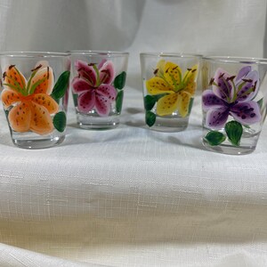 May include: Set of four clear glass shot glasses with hand-painted lily flowers. Each glass features a different colored lily: orange, pink, yellow, and purple.