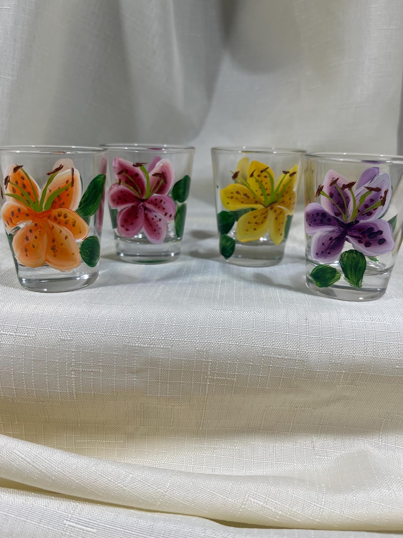 May include: Set of four clear glass shot glasses with hand-painted lily flowers. Each glass features a different colored lily: orange, pink, yellow, and purple.