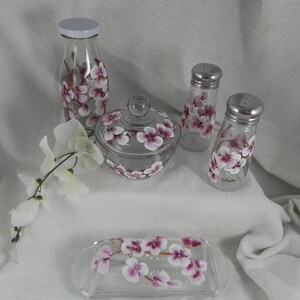 May include: Set of five hand-painted glass kitchen containers with pink and white floral designs. The set includes a tall bottle, a sugar bowl with lid, two salt and pepper shakers, and a butter dish.