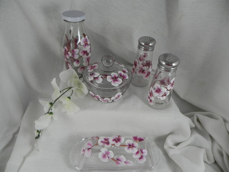 May include: Set of five hand-painted glass kitchen containers with pink and white floral designs. The set includes a tall bottle, a sugar bowl with lid, two salt and pepper shakers, and a butter dish.