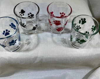 Puede incluir: Cuatro vasos de chupito transparentes con diseños de huellas de patas en azul, negro, rojo y verde. Los vasos están colocados sobre una superficie blanca, mostrando las coloridas huellas.
