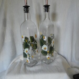 May include: Two clear glass bottles with white and yellow floral designs. The bottles have stainless steel spouts and black tops. The bottles are on a white surface.