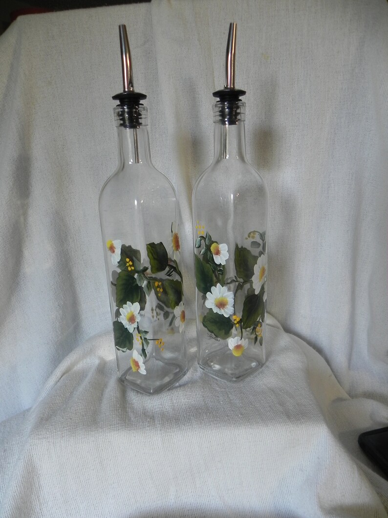 May include: Two clear glass bottles with white and yellow floral designs. The bottles have stainless steel spouts and black tops. The bottles are on a white surface.