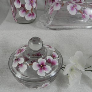 May include: A clear glass butter dish with a lid, hand-painted with pink and white flowers. The dish is sitting on a white surface.