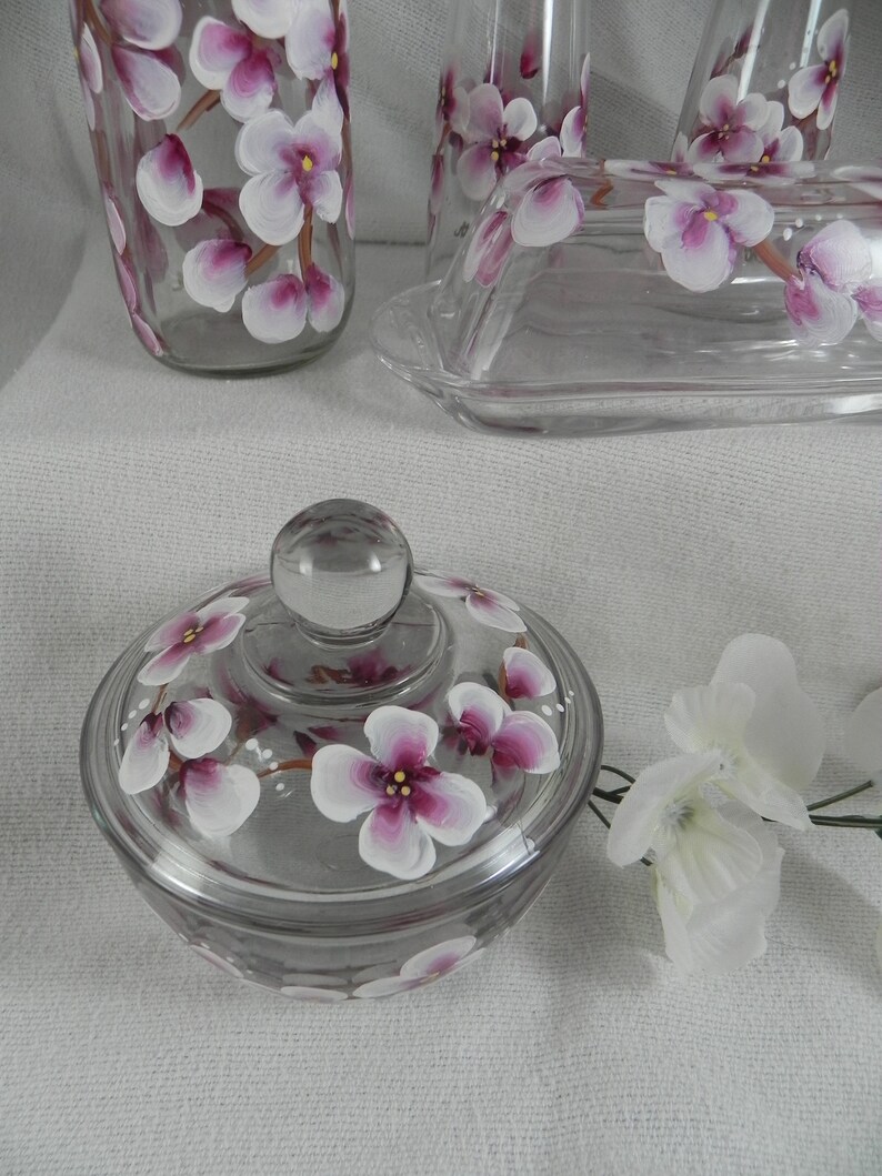 May include: A clear glass butter dish with a lid, hand-painted with pink and white flowers. The dish is sitting on a white surface.