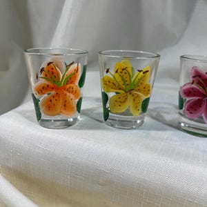 May include: Three clear glass shot glasses with hand-painted lily flowers. The glasses feature orange, yellow, and pink lilies with green leaves.