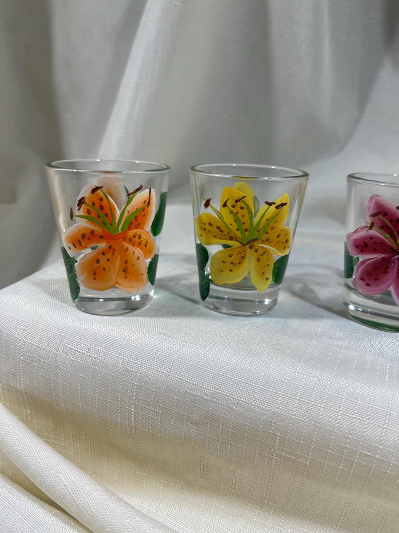 May include: Three clear glass shot glasses with hand-painted lily flowers. The glasses feature orange, yellow, and pink lilies with green leaves.