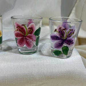 May include: Set of three clear glass shot glasses with hand-painted floral designs. Each glass features a different colored lily flower with green leaves.