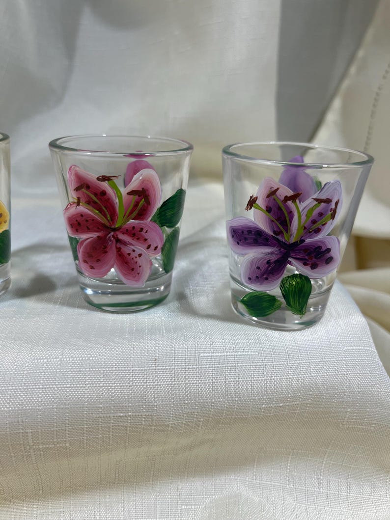 May include: Set of three clear glass shot glasses with hand-painted floral designs. Each glass features a different colored lily flower with green leaves.