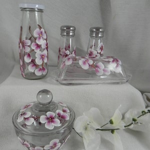 May include: Set of hand-painted glass kitchenware featuring a floral design with pink and white flowers. The set includes a milk bottle, two salt and pepper shakers, a butter dish, and a sugar bowl with lid.