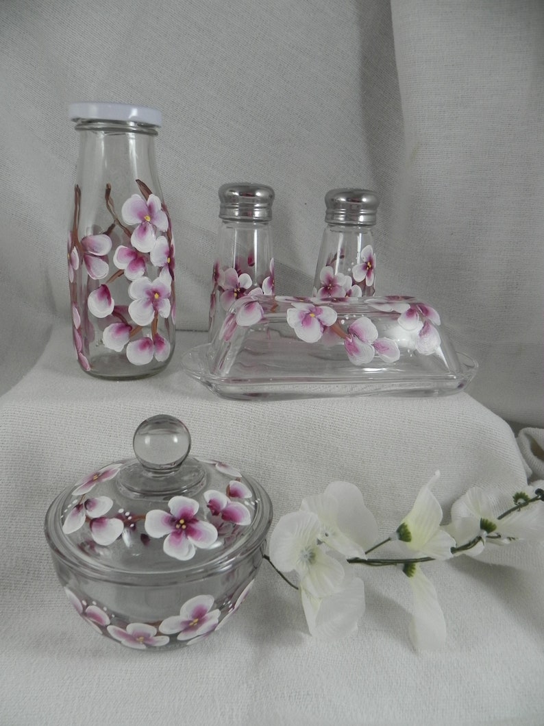 May include: Set of hand-painted glass kitchenware featuring a floral design with pink and white flowers. The set includes a milk bottle, two salt and pepper shakers, a butter dish, and a sugar bowl with lid.