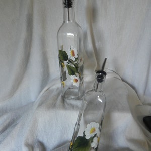 May include: Two clear glass bottles with white daisy and green leaf designs. The bottles have black pour spouts and are on a white fabric background.