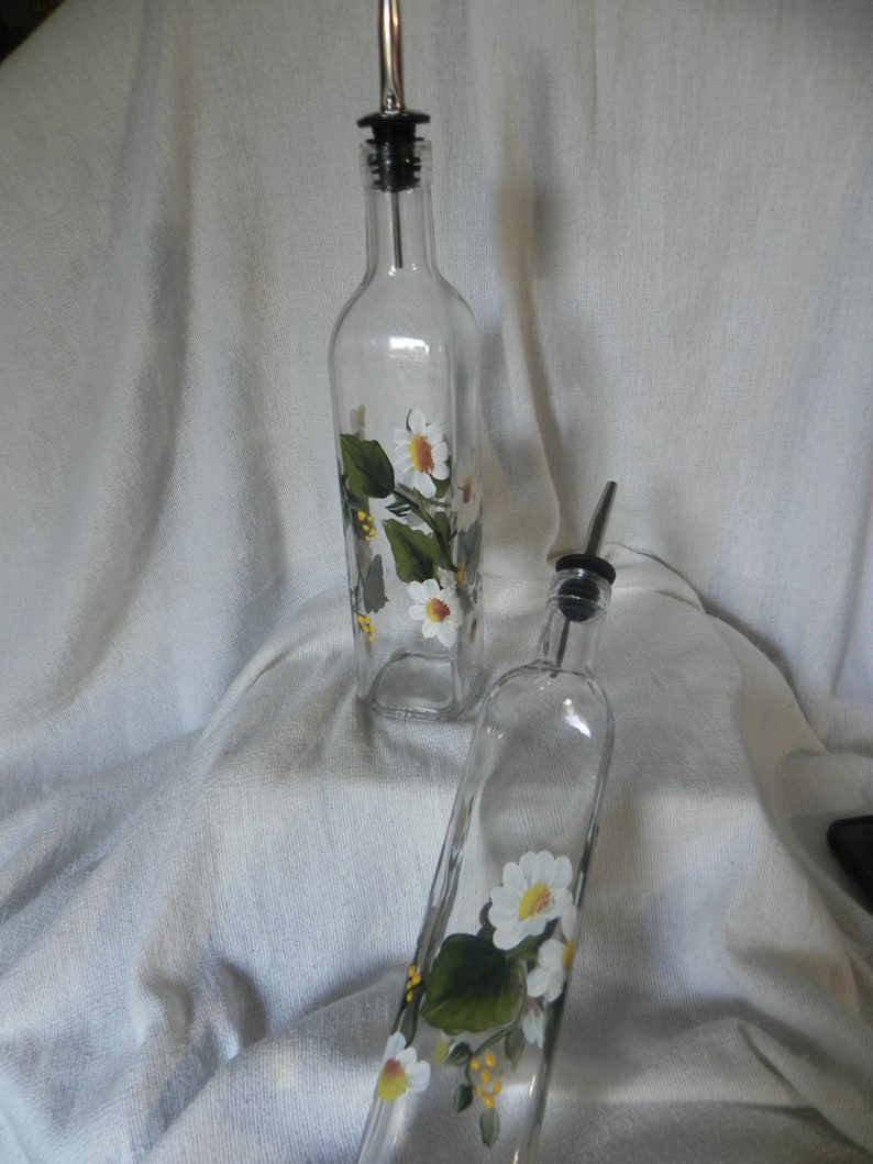 May include: Two clear glass bottles with white daisy and green leaf designs. The bottles have black pour spouts and are on a white fabric background.