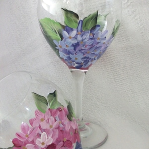 May include: Two hand-painted wine glasses with floral designs. The glasses feature a cluster of purple and pink flowers with green leaves.