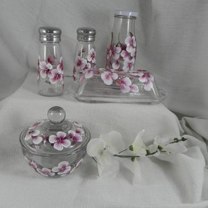 May include: Set of four glass kitchen containers with hand-painted pink and white floral designs. The set includes a butter dish, two salt and pepper shakers, and a sugar bowl with a lid.