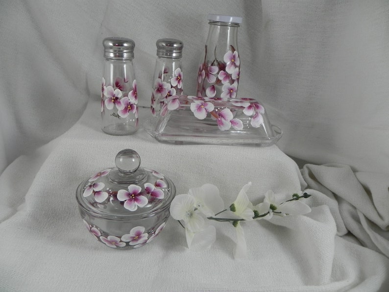 May include: Set of four glass kitchen containers with hand-painted pink and white floral designs. The set includes a butter dish, two salt and pepper shakers, and a sugar bowl with a lid.