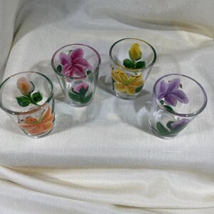May include: Set of four clear glass shot glasses with hand-painted floral designs. Each glass features a different flower in shades of pink, yellow, orange, and purple.