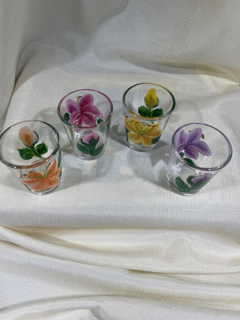 May include: Set of four clear glass shot glasses with hand-painted floral designs. Each glass features a different flower in shades of pink, yellow, orange, and purple.