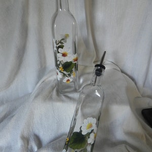 May include: Two clear glass bottles with black pour spouts, hand-painted with white daisies and green leaves. The bottles are on a white fabric background.