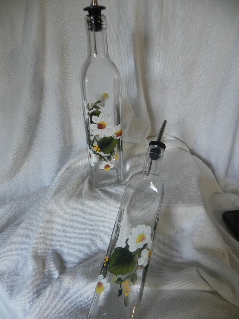 May include: Two clear glass bottles with black pour spouts, hand-painted with white daisies and green leaves. The bottles are on a white fabric background.