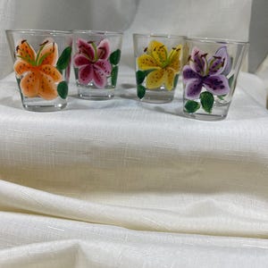 May include: Four clear glass shot glasses with hand-painted lily flowers. Each glass features a different colored lily: orange, pink, yellow, and purple.