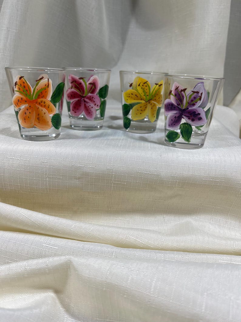 May include: Four clear glass shot glasses with hand-painted lily flowers. Each glass features a different colored lily: orange, pink, yellow, and purple.
