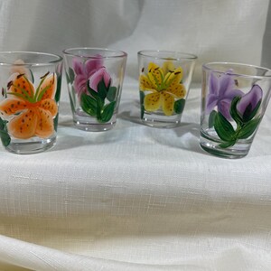 May include: Four clear glass shot glasses with hand-painted floral designs. Each glass features a different flower: orange lily, pink rose, yellow lily, and purple flower.