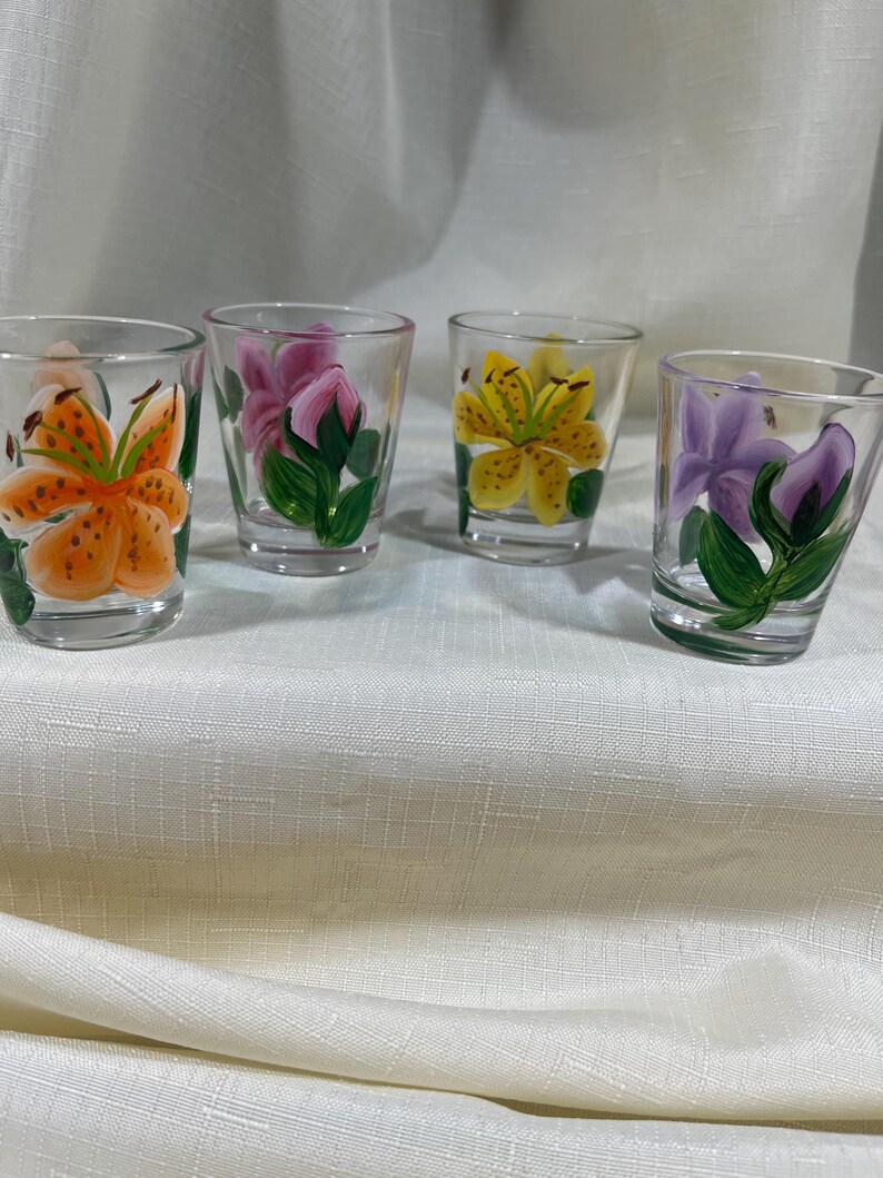May include: Four clear glass shot glasses with hand-painted floral designs. Each glass features a different flower: orange lily, pink rose, yellow lily, and purple flower.