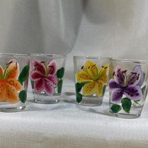 May include: Set of four clear glass shot glasses with hand-painted lily flower designs. Each glass features a different colored lily flower: orange, pink, yellow, and purple.