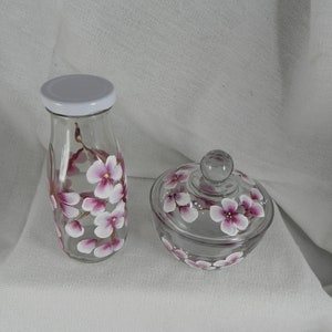 May include: A set of two glass jars with hand-painted pink cherry blossoms. The larger jar is a milk bottle shape with a white lid. The smaller jar is a round jar with a lid. Both jars have a floral design.