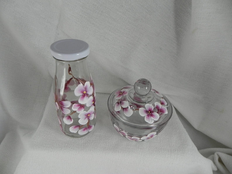 May include: A set of two glass jars with hand-painted pink cherry blossoms. The larger jar is a milk bottle shape with a white lid. The smaller jar is a round jar with a lid. Both jars have a floral design.