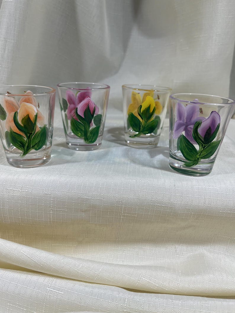 May include: Four clear glass shot glasses with hand-painted floral designs. Each glass features a different colored flower: peach, pink, yellow, and purple.