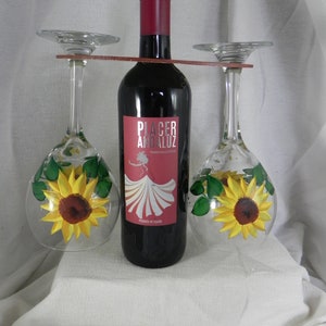 Puede incluir: Un porta botellas de vino con dos copas de vino. El porta botellas está hecho de madera y tiene un acabado marrón. Las copas de vino son de vidrio transparente con girasoles pintados y hojas verdes. La botella de vino es roja y tiene la etiqueta "Placer Andaluz Tempranillo Crianza".